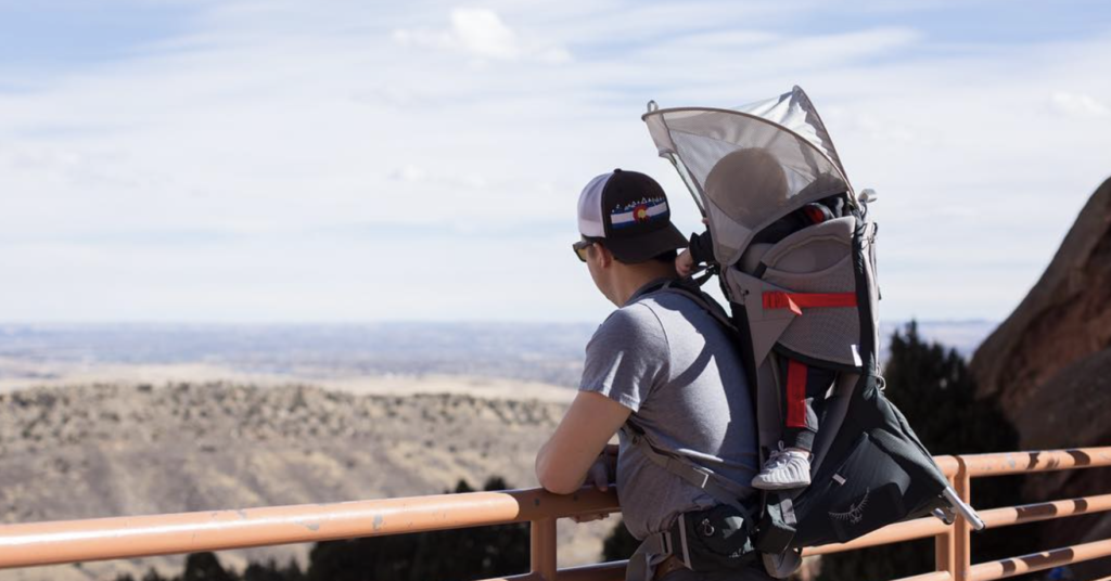 A man with a baby in a backpack embarks on outdoor adventures for kids near Denver, taking in the breathtaking views of Red Rocks Amphitheatre.