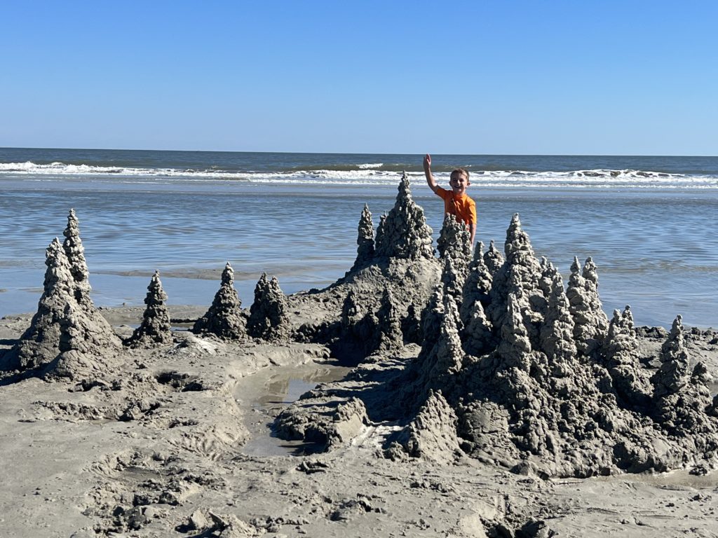 A boy standing in front of a sand castle on the beach.