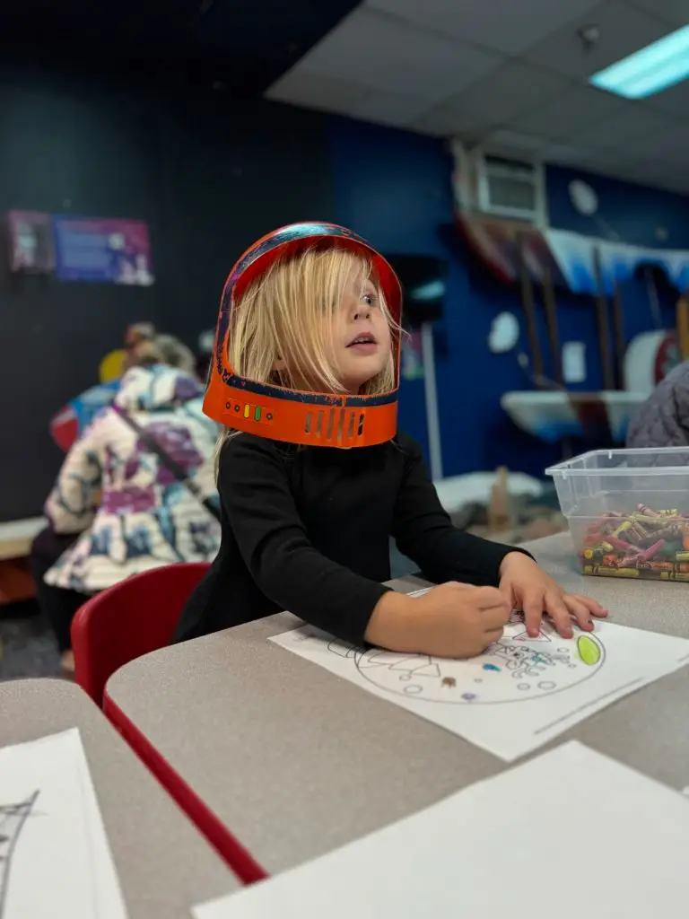 A young girl wearing a space helmet at a table at Wings Over The Rockies With Kids