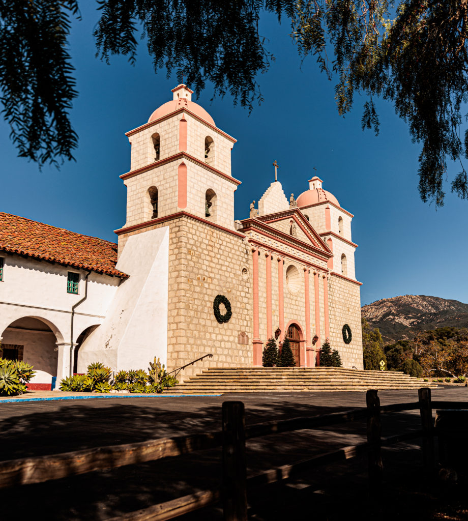 A church with two towers and a wooden fence. Old Mission Santa Barbara with kids.