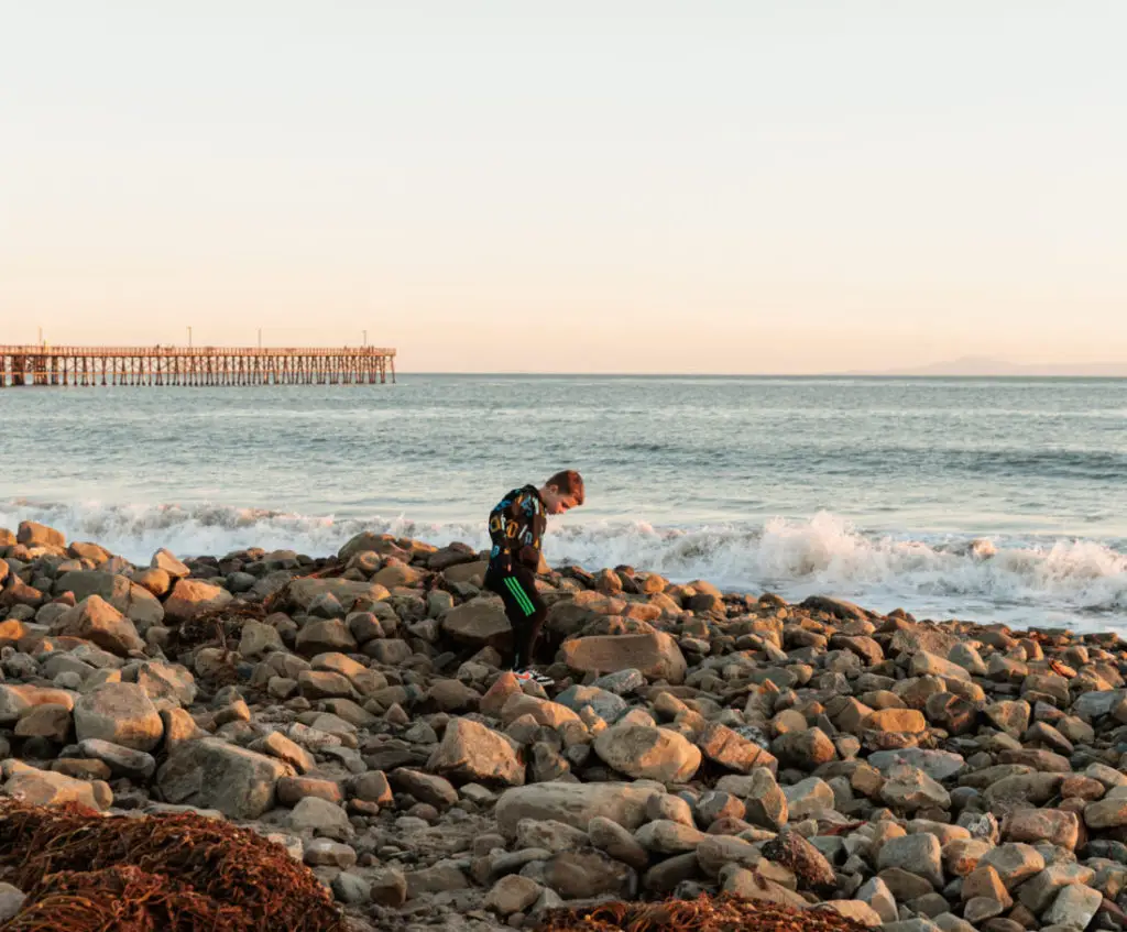 A boy is standing on a rocky beach near a pier. Beaches in Santa Barbara With Kids