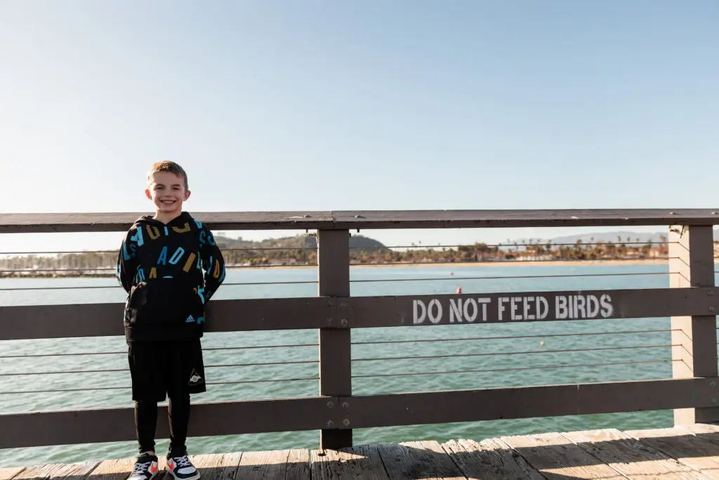 A boy standing on a wooden bridge with a sign that says don't feed the birds. Stearns Wharf with kids.