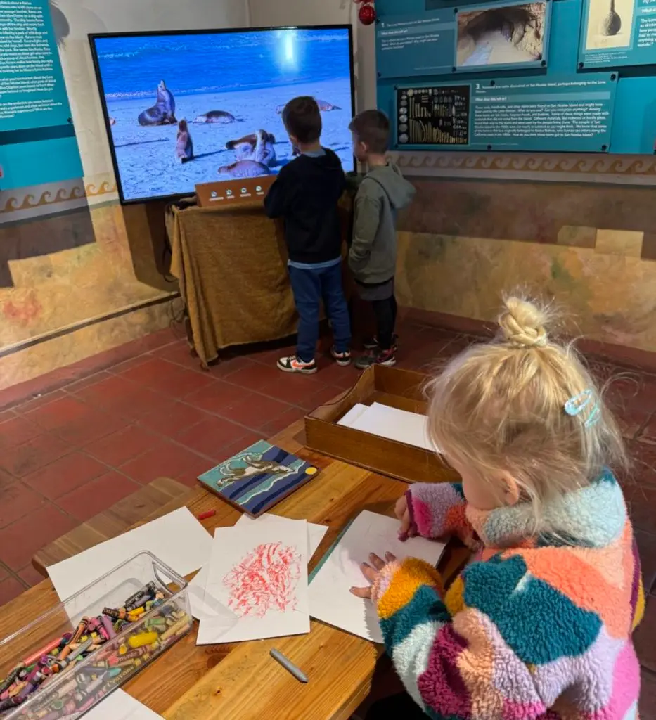 A group of children drawing on a table in front of a tv. Old Mission Santa Barbara with kids.