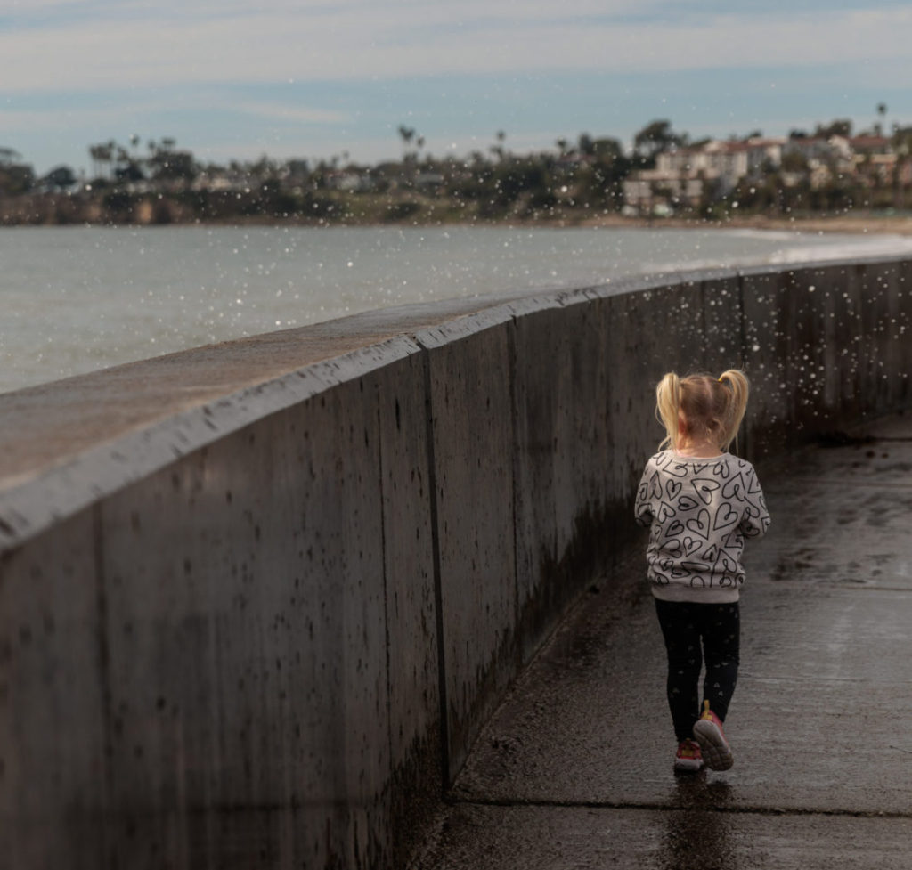 A little girl is walking along a concrete wall near the ocean at the Santa Barbara Harbor Breakwater Wall. 