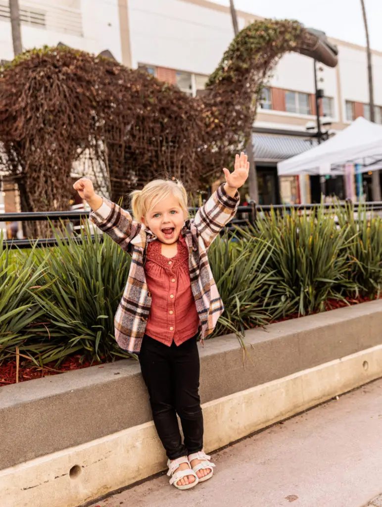 A little girl is standing in front of a statue of a dinosaur. 3rd Street Promenade with kids