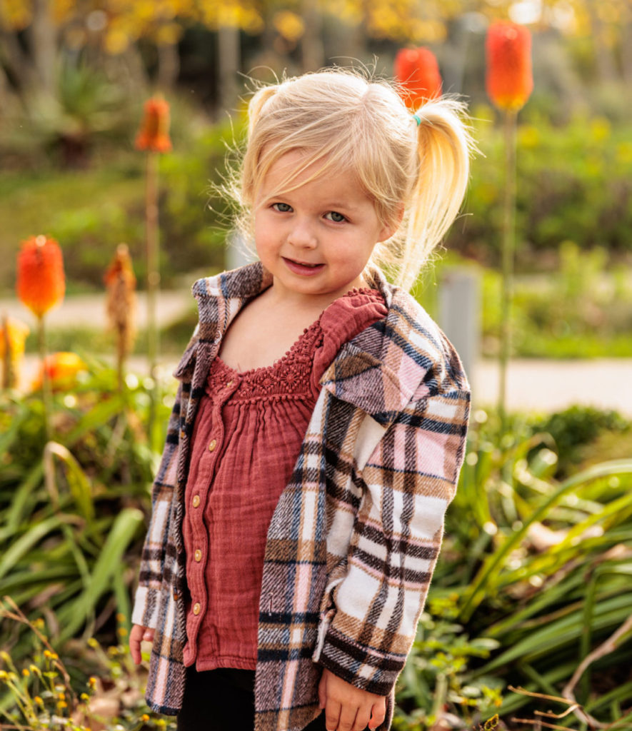 A young girl with blonde hair, wearing a plaid jacket and red top, stands in a garden with orange flowers in the background.  Tongva Park, Santa Monica Playground