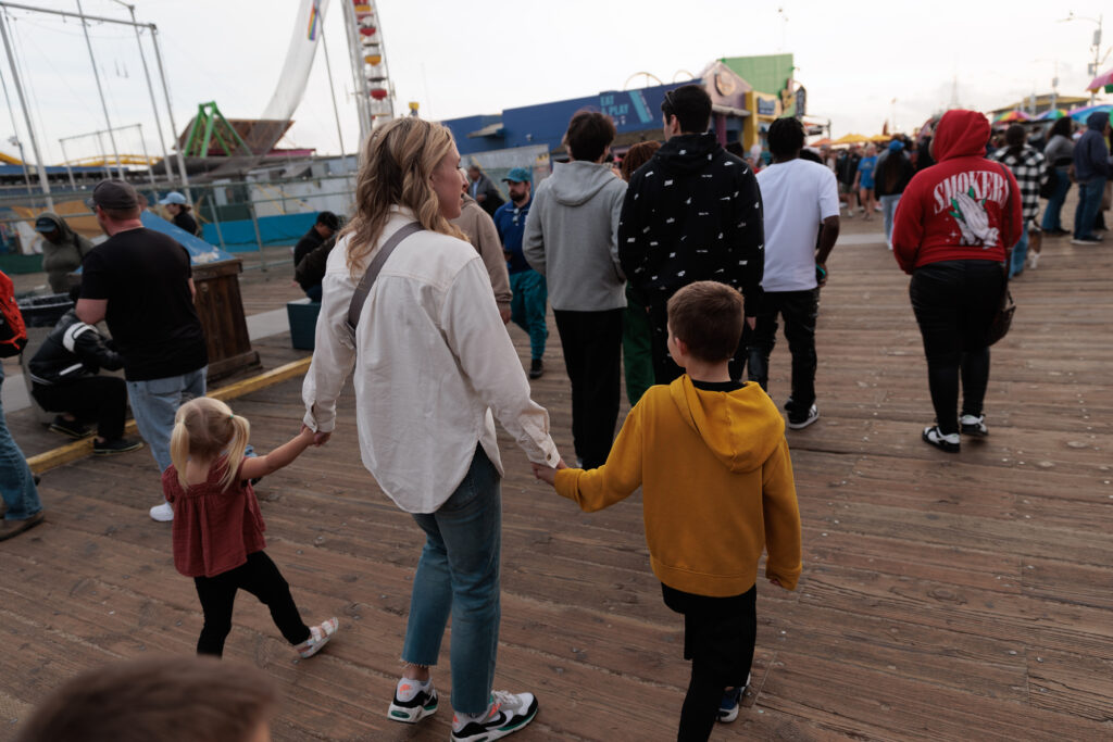 A woman walks with two children holding hands on a wooden boardwalk, surrounded by people. The background includes amusement park rides and structures. Santa Monica Pier with Kids