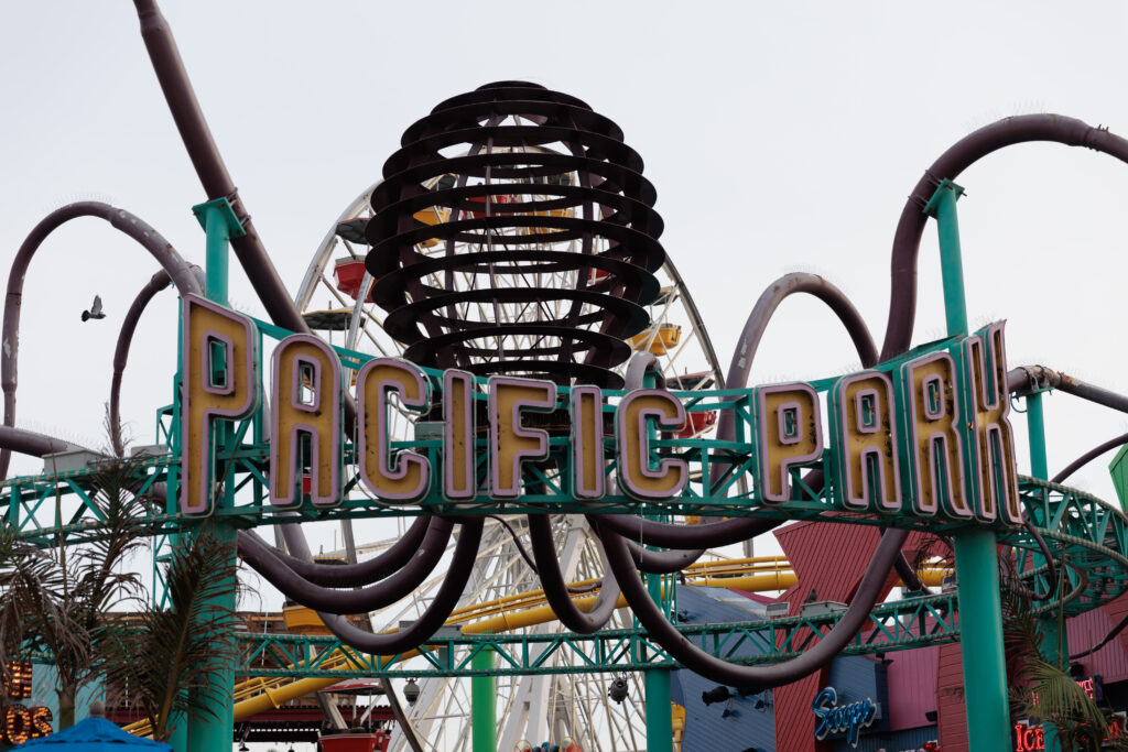 Close-up view of the entrance to Pacific Park, with a large Ferris wheel partially visible in the background. The sign has bold letters and is adorned with roller coaster tracks and other rides. Santa Monica Pier with Kids