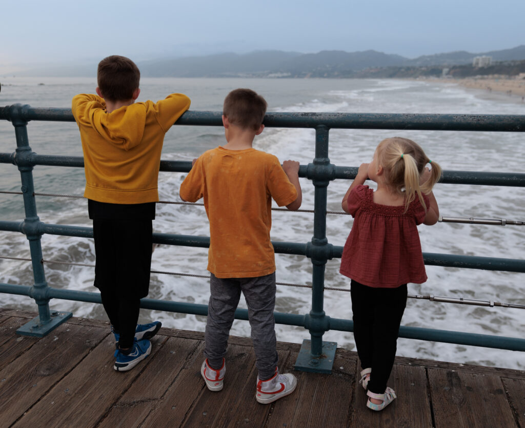 Three children stand on a pier facing the ocean, with their hands on the railings, looking at the waves and the distant shoreline under a cloudy sky.  Santa Monica Pier with Kids