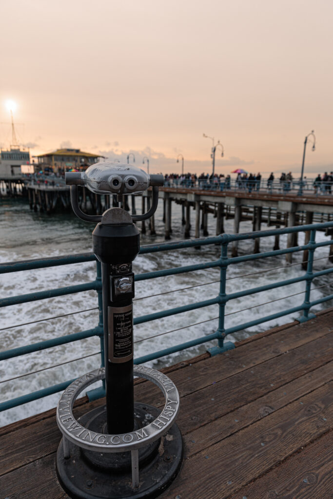 A coin-operated binocular viewer on a wooden pier overlooks the ocean, with a distant building and people gathered near the railing at sunset. Santa Monica Pier with Kids