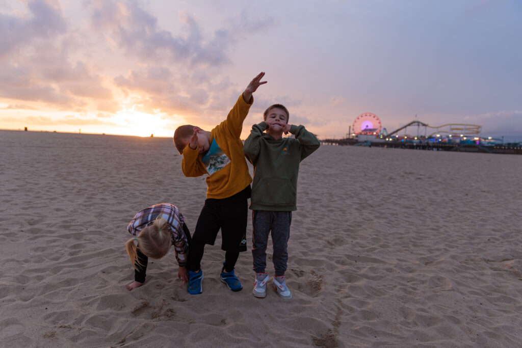 Three children pose on a sandy beach at sunset with a lit-up Ferris wheel in the background; one child bends down, while the others strike playful poses, one dabbing and the other with hands up. Santa Monica Pier with Kids