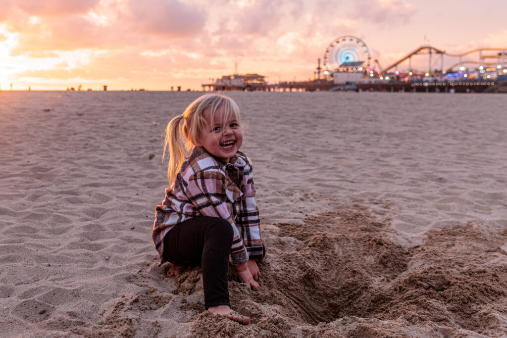 A young child with blond hair and a plaid jacket smiles while digging in the sand on a beach at sunset. An amusement park with a Ferris wheel is visible in the background. Santa Monica Pier with Kids