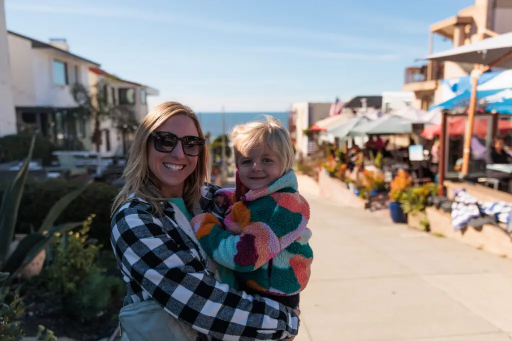 A woman wearing sunglasses holds a young child in a colorful jacket. They are on a sunny outdoor path with shops and cafes in the background.  A day in Manhattan Beach with kids.