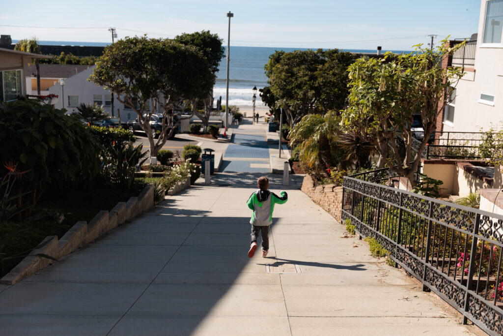 A child in a green and white shirt runs down a paved residential street towards the ocean on a sunny day, with trees and houses lining the street.  A day in Manhattan Beach with kids.