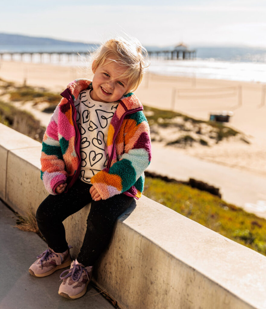 A young child wearing a colorful jacket and heart-patterned shirt smiles while sitting on a concrete ledge at the beach. The sunny beach and a pier are visible in the background. A day in Manhattan Beach with kids.