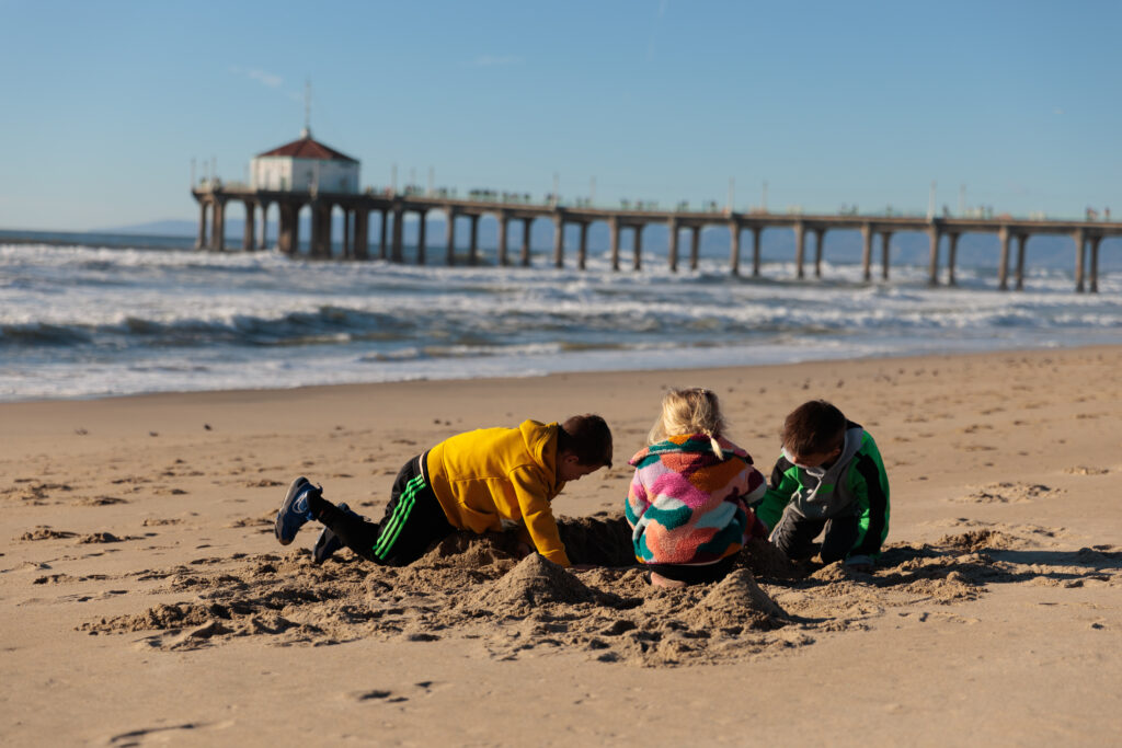 Three children play in the sand on a beach with a long pier and ocean waves in the background under a clear blue sky.  A day in Manhattan Beach with kids.