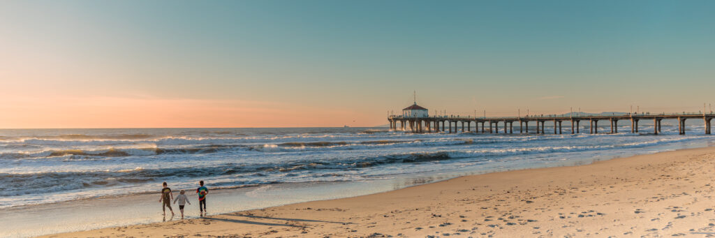 Two people walk along the shoreline near a pier during sunset, with waves crashing onto the sandy beach. A day in Manhattan Beach with kids.