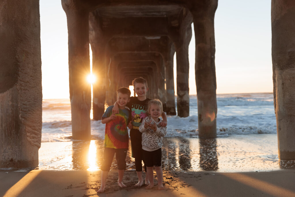 Three children smiling and posing under a pier at the beach during sunset, with waves in the background and the sun casting a warm glow.  A day in Manhattan Beach with kids.