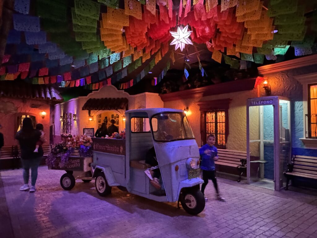 A colorful scene featuring a blue three-wheeled cart filled with flowers, vibrant paper decorations overhead, a star-shaped light, people walking, and a vintage telephone booth on the right. Casa Bonita with the family. 