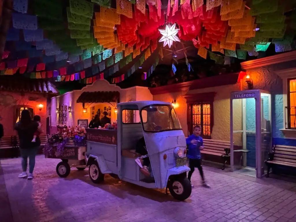 A colorful scene featuring a blue three-wheeled cart filled with flowers, vibrant paper decorations overhead, a star-shaped light, people walking, and a vintage telephone booth on the right. Casa Bonita with the family. 