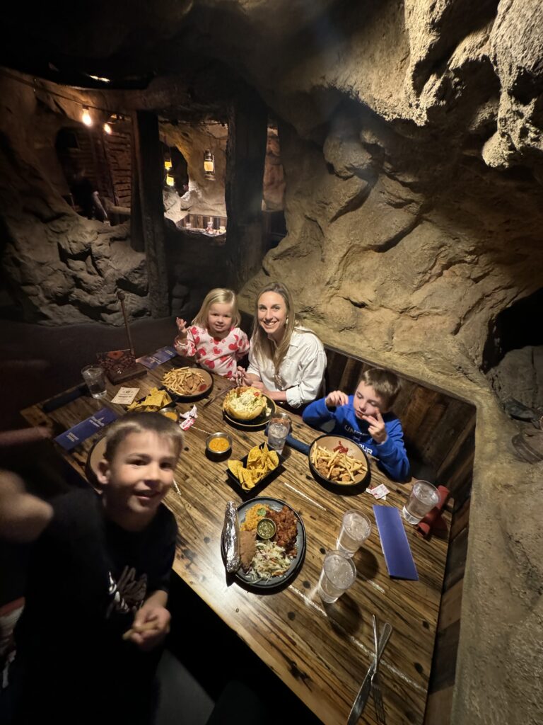 A woman and three children sit at a table in a dimly lit, cave-themed restaurant, smiling and enjoying their meals. Casa Bonita with the family. 