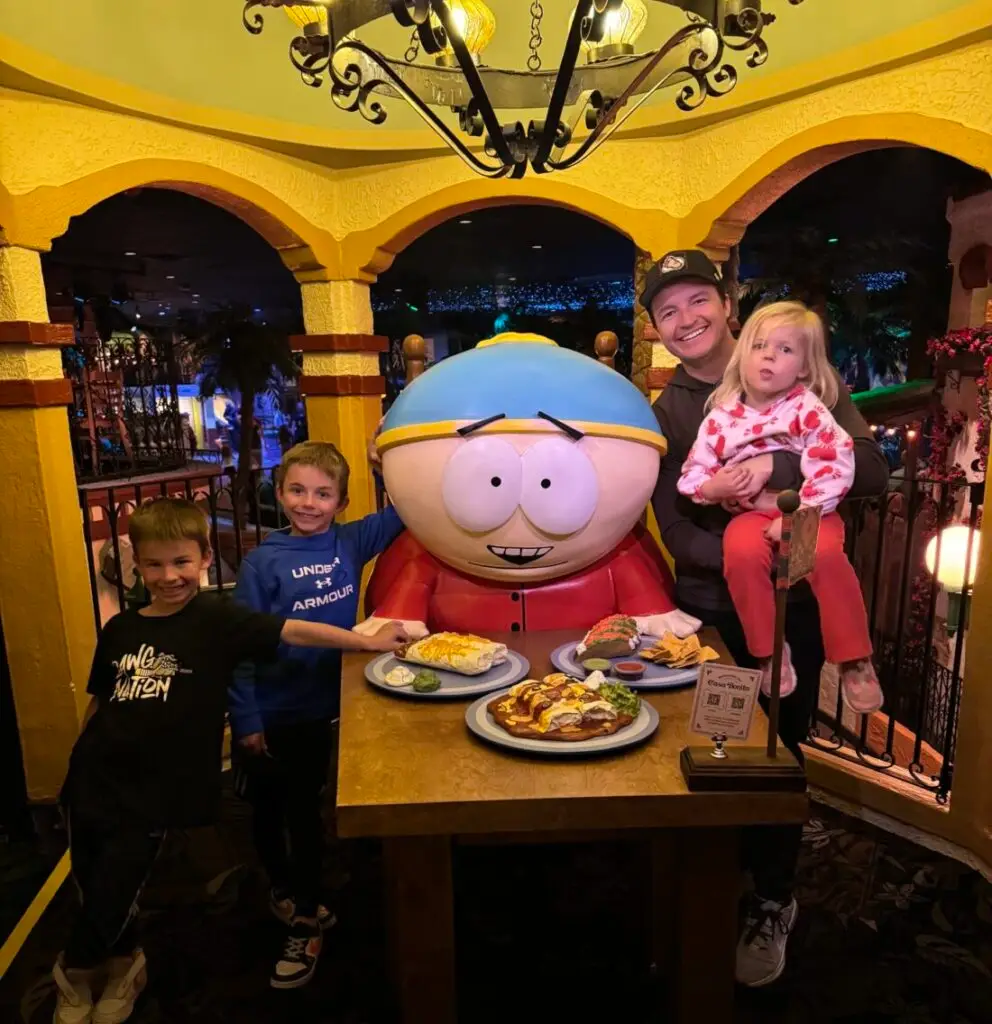 A person and three children pose with a large statue of a cartoon character at a table with plates of food on it, under a chandelier in a themed restaurant setting. Casa Bonita with the family. 