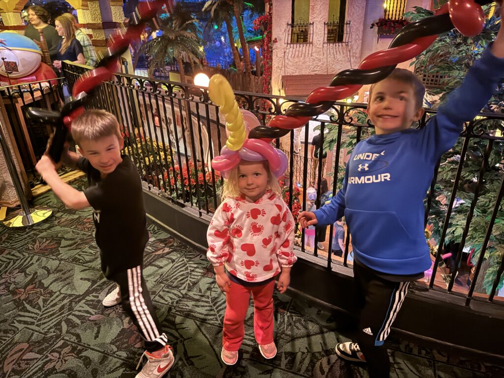 Three children are indoors. Two boys with balloon swords stand on either side of a girl wearing a balloon hat. They are on a decorative carpeted floor with plants in the background. Casa Bonita with the family. 