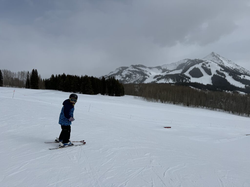 A person wearing a blue jacket and black helmet is skiing on a snowy slope with mountains and trees visible in the background. The sky is overcast. Crested Butte family vacation.