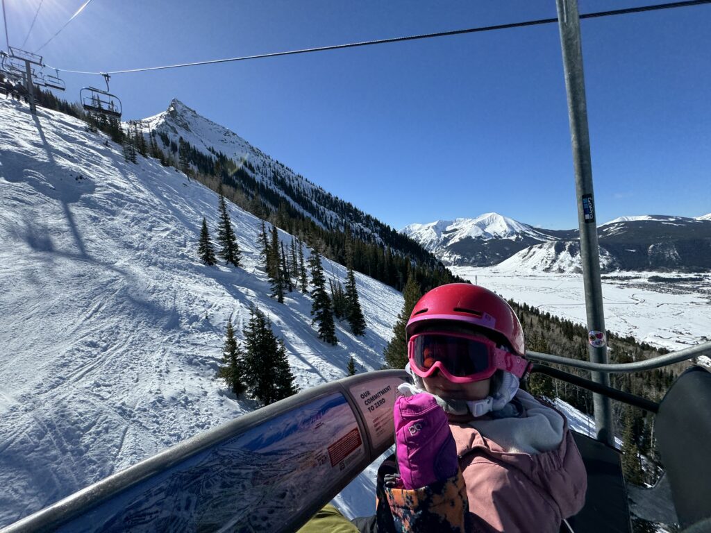 A person wearing a pink helmet and goggles rides a ski lift overlooking snowy mountain slopes and distant peaks on a clear day. Crested Butte family vacation.