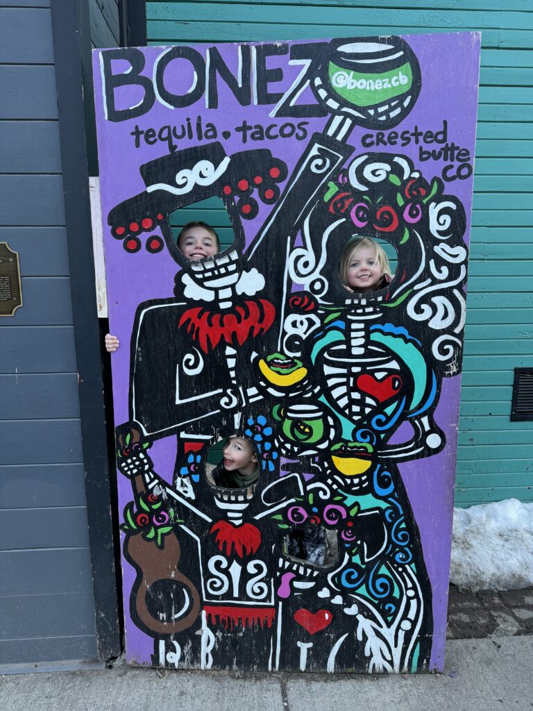 Three children pose with their faces in cut-outs of a painted board featuring festive skeletons holding food and drinks. The board reads "Bonez" and "tequila - tacos. Crested Butte family vacation.