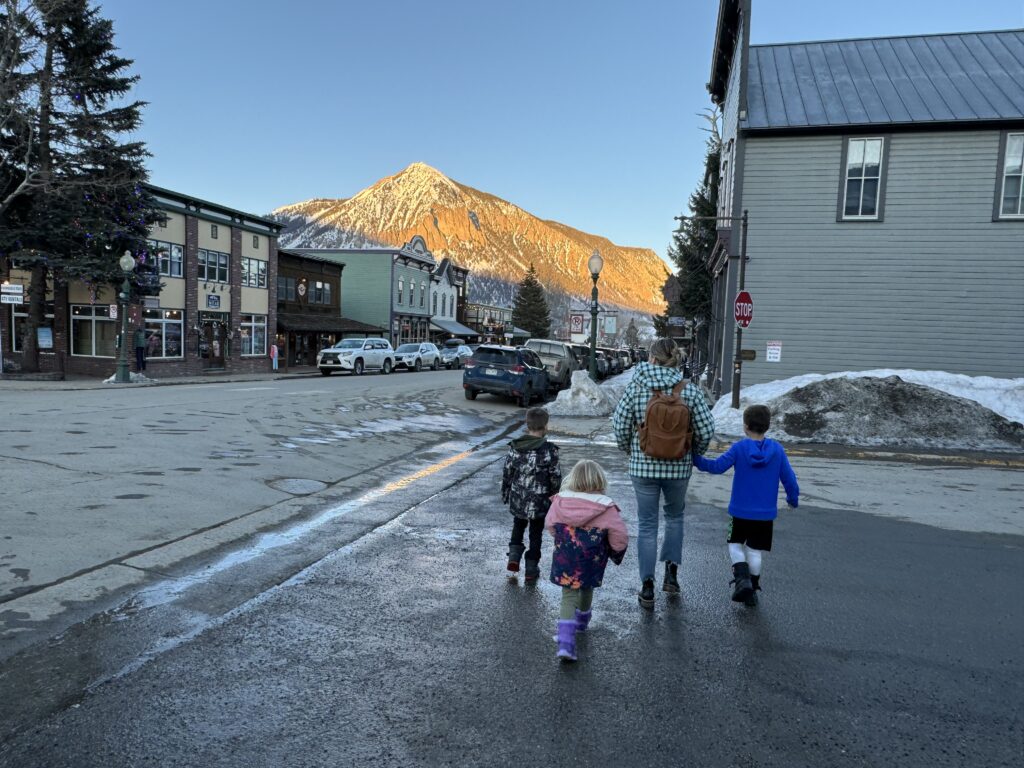 Four people walking down a street in a small town with a mountain in the background on a clear day. Snow is visible on the sides of the road. Crested Butte family vacation.