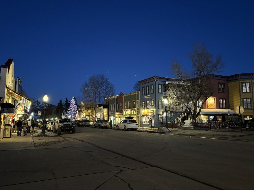 A small town street at dusk, with lit buildings and Christmas lights on a tree, cars parked along the sides, and a few people walking. Crested Butte family vacation.