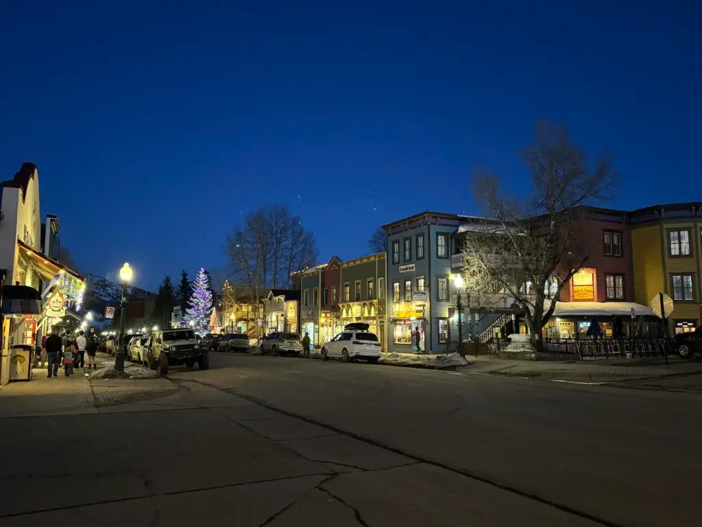 A small town street at dusk, with lit buildings and Christmas lights on a tree, cars parked along the sides, and a few people walking. Crested Butte family vacation.