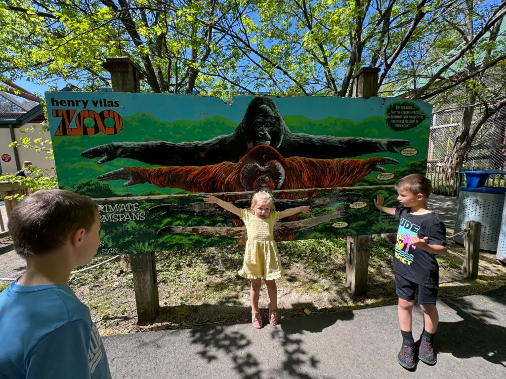 Three children stand in front of a gorilla and orangutan sign at Henry Vilas Zoo, each posing with their arms spread out to match the gorilla's outstretched arms depicted on the sign. Free zoo in Madison