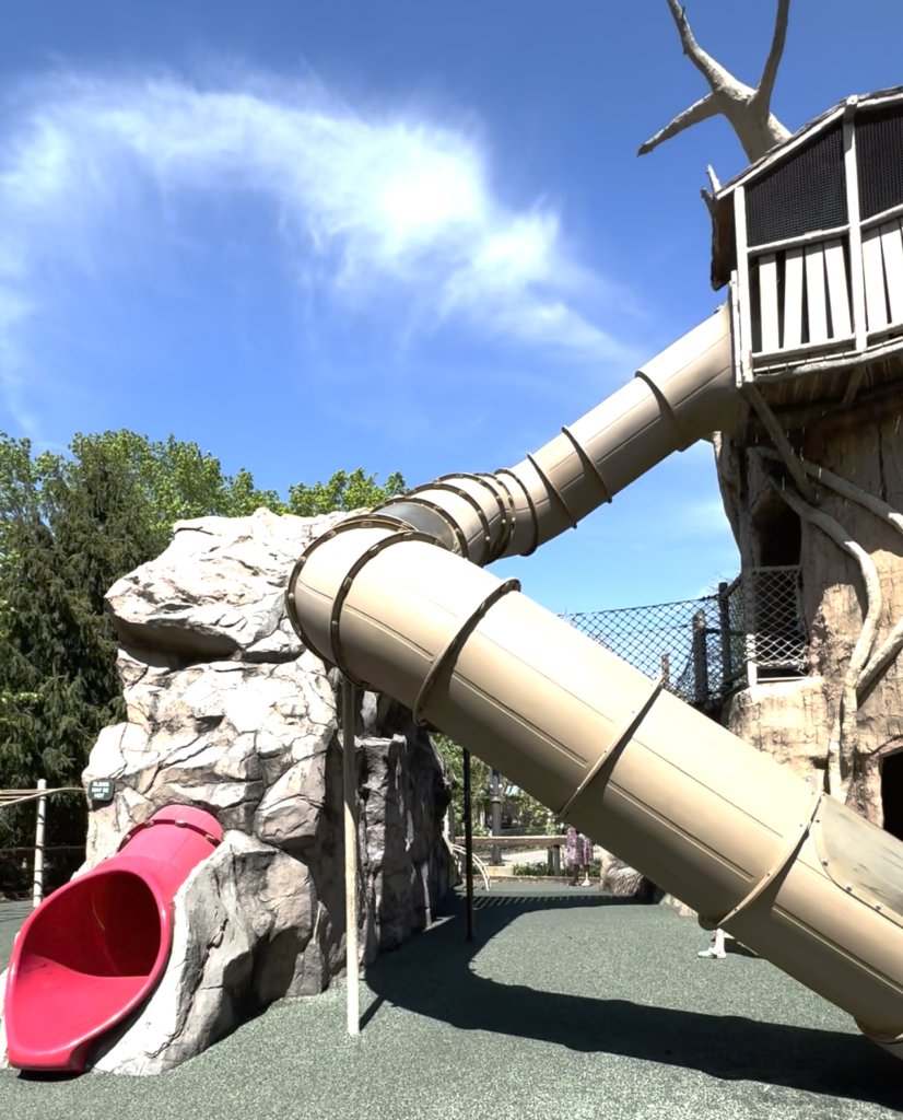 A playground with a tall wooden structure, featuring two tube slides, beige and red, against a clear blue sky.  Free zoo in Madison