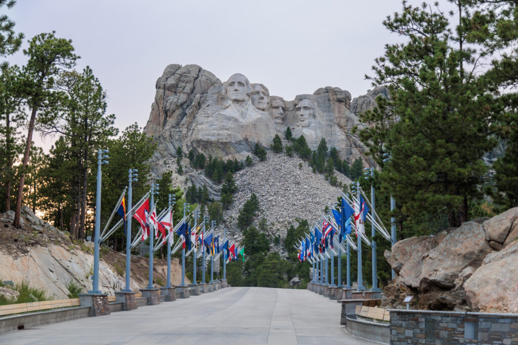 Mount Rushmore National Memorial, showing the carved faces of four U.S. presidents, with an avenue of international flags leading up to it, surrounded by trees and rocky terrain. Mount Rushmore with kids.