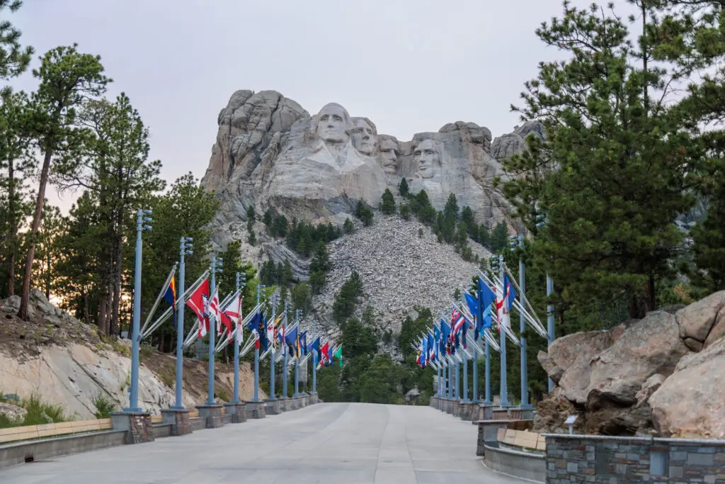 Mount Rushmore National Memorial, showing the carved faces of four U.S. presidents, with an avenue of international flags leading up to it, surrounded by trees and rocky terrain. Mount Rushmore with kids.