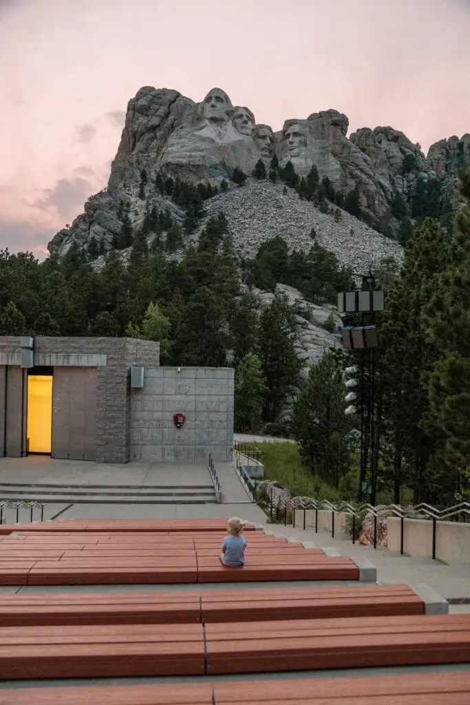 A child sits alone on benches facing Mount Rushmore, with the iconic carved presidential faces visible in the mountain in the background.  Mount Rushmore with kids