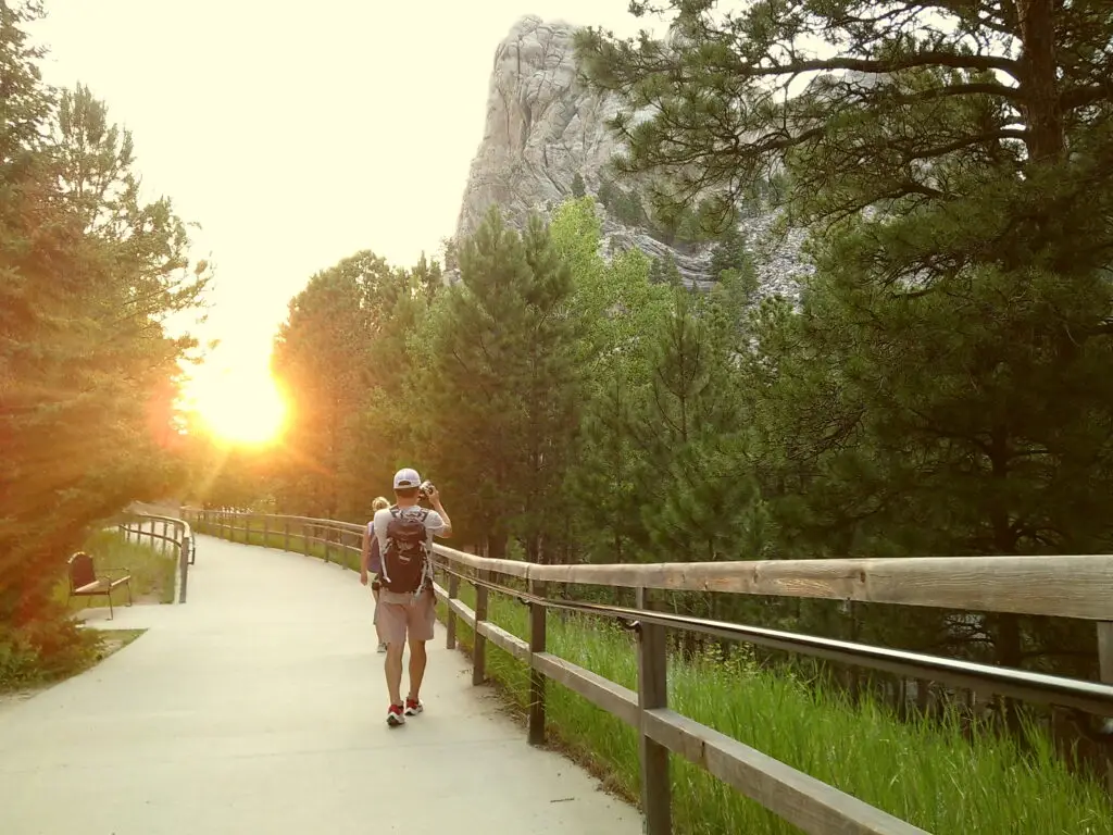 A person walks along a paved trail surrounded by trees with a large rock formation in the background during sunset.  Mount Rushmore with kids