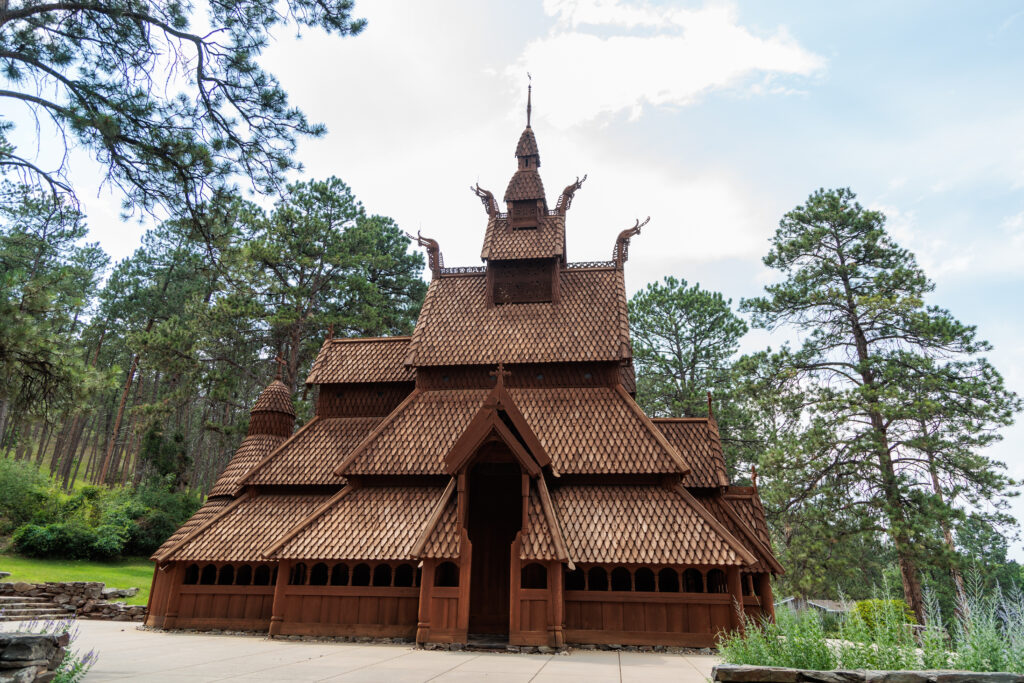 A wooden stave church with intricate carvings stands amidst tall pine trees under a partly cloudy sky. Free things to do in Rapid City with kids.