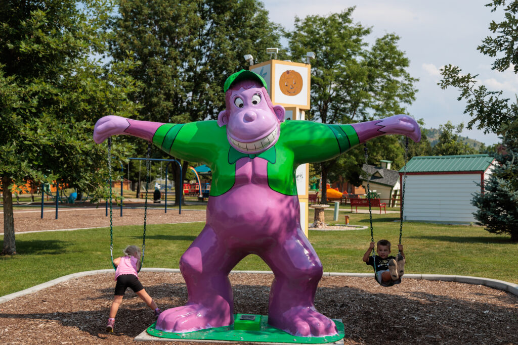 Two children are using a swing set that is built into a large, colorful statue of a cartoonish gorilla in a park. Trees and playground structures are visible in the background.  Free things to do in Rapid City with kids.