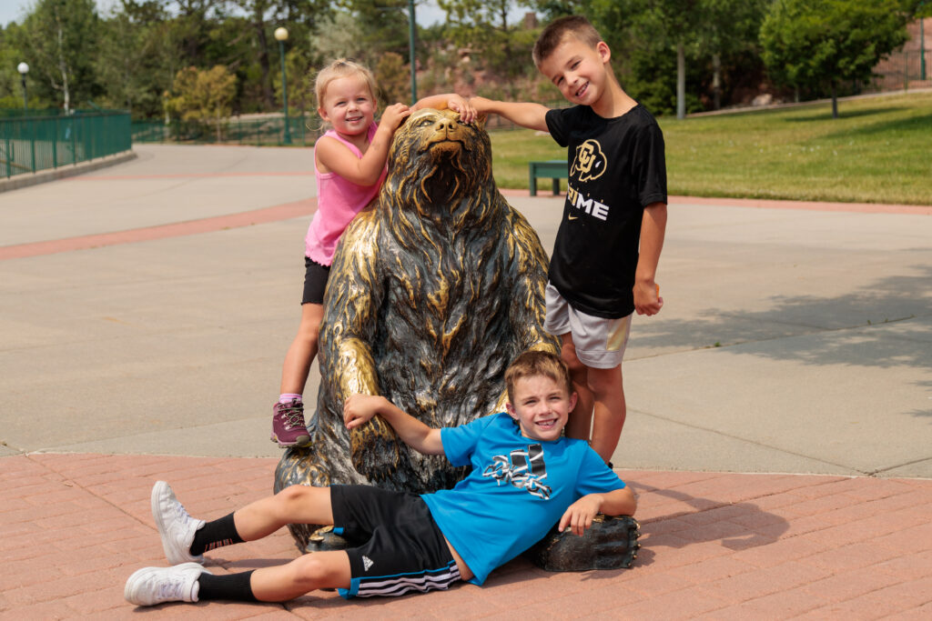 Three smiling children posing with a bear statue outdoors. One child lies on the ground while the other two stand beside and on the statue, placing their hands on it. Things to do in South Dakota with kids, Bear Country USA.