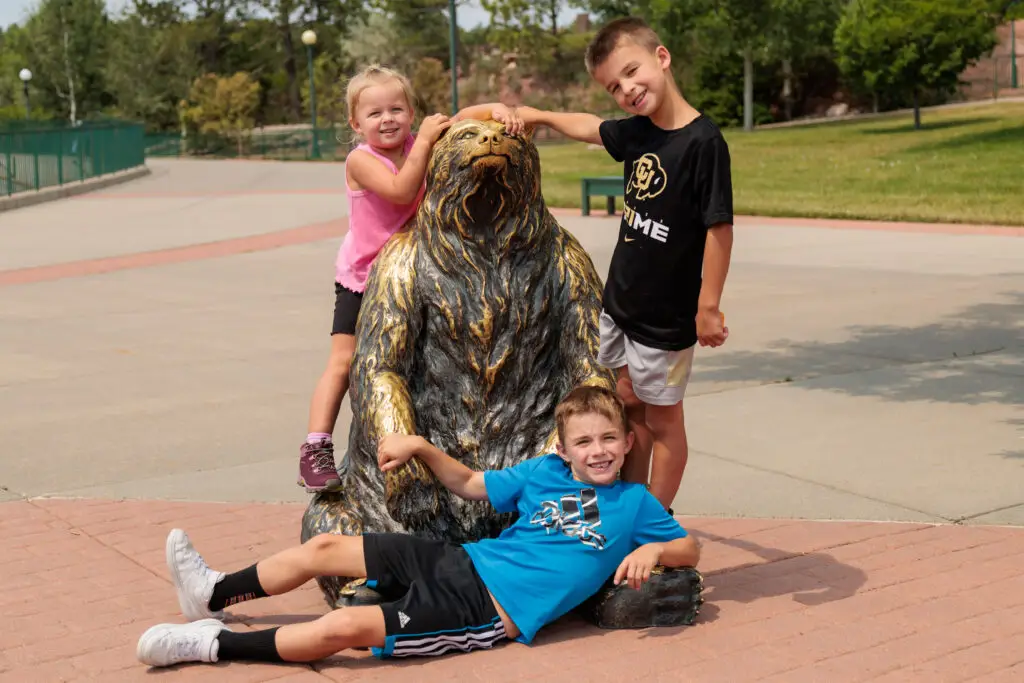 Three smiling children posing with a bear statue outdoors. One child lies on the ground while the other two stand beside and on the statue, placing their hands on it. Things to do in South Dakota with kids, Bear Country USA.