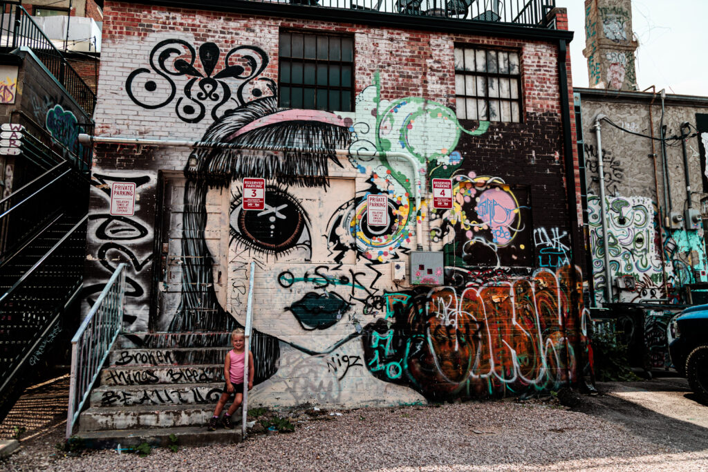 A child stands on stairs in front of a brick wall covered with colorful graffiti art, including a large mural of a girl's face with big eyes. Free things to do in Rapid City with kids.