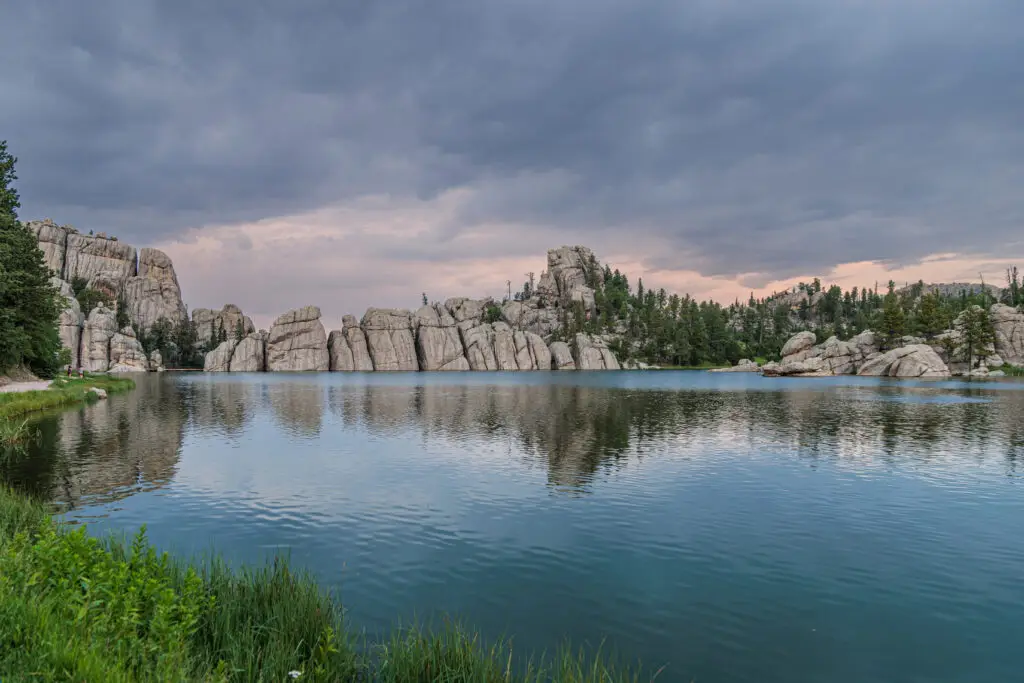 A serene lake surrounded by rocky cliffs and evergreen trees under a cloudy sky, with still water reflecting the landscape. Custer State Park with kids.