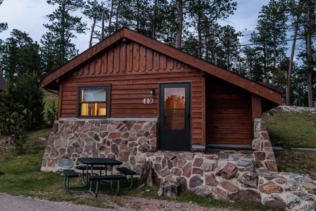 A small, single-story wooden cabin with a stone exterior and the number "410" on the wall. It has a well-lit window and door, surrounded by trees, and features an outdoor seating area with a picnic table. Custer State Park with kids.