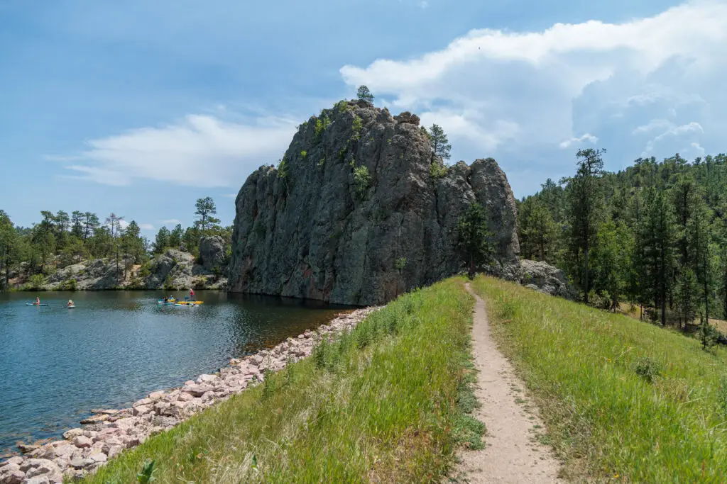 A dirt path runs alongside a scenic lake with rocky cliffs and pine trees under a partly cloudy sky. Custer State Park with kids.