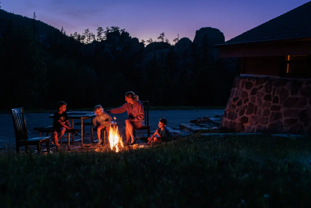 A group of people sit around a campfire in an outdoor setting at dusk, near a stone building and trees in the background. Custer State Park with kids.