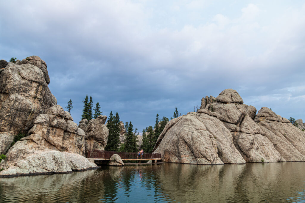 Rock formations surround a small bridge over a calm lake, with pine trees and a cloudy sky in the background. Two people are walking on the bridge. Custer State Park with kids.