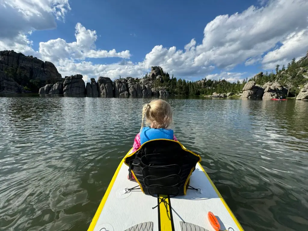 A child with blonde hair, dressed in a blue life jacket, sits on a paddleboard facing a lake surrounded by rocky cliffs and forested hills under a partly cloudy sky.  Custer State Park with kids.